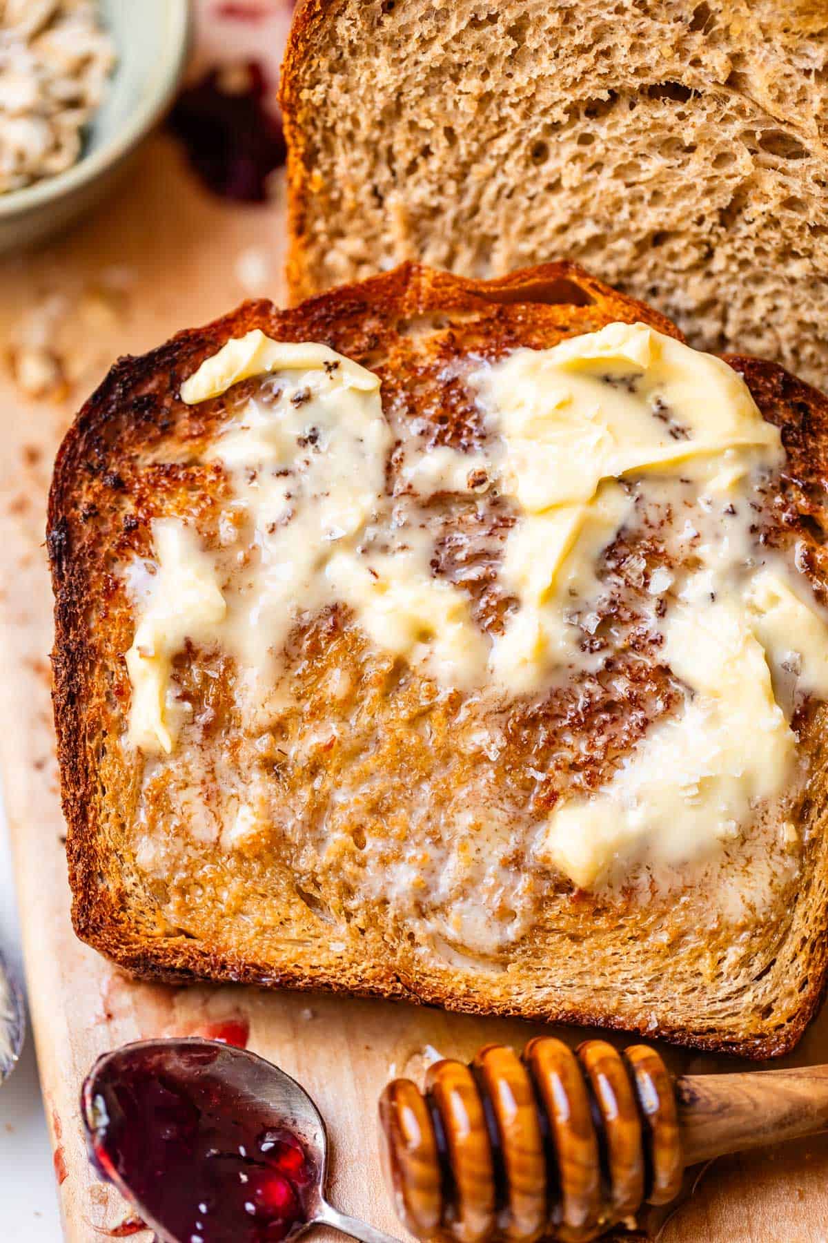 A slice of toasted bread topped with melting butter, next to a loaf of bread, a honey dipper, and a spoon with dark fruit jam on a wooden board.