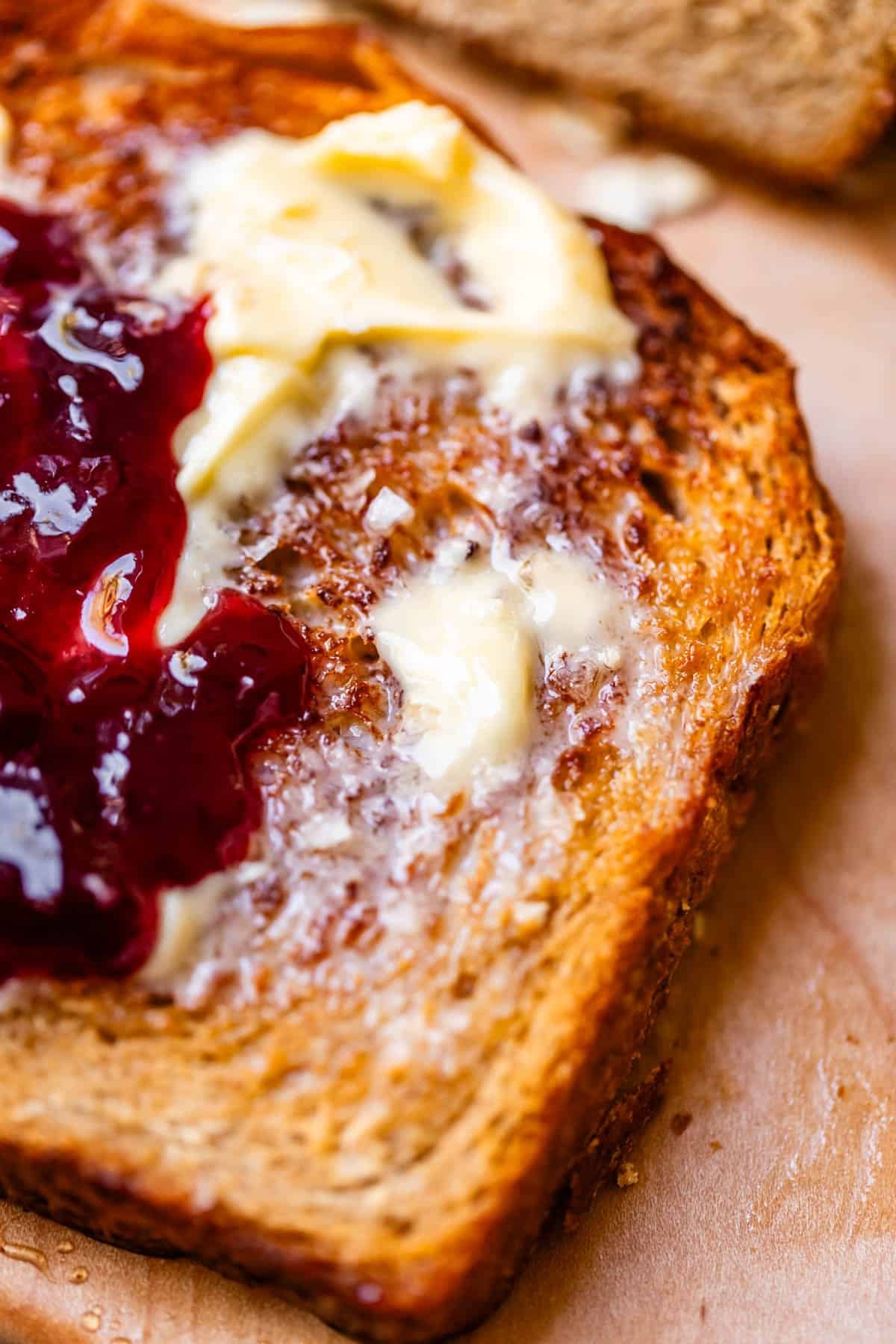 A close-up of toasted bread spread with melting butter and red fruit jam, placed on a wooden surface.