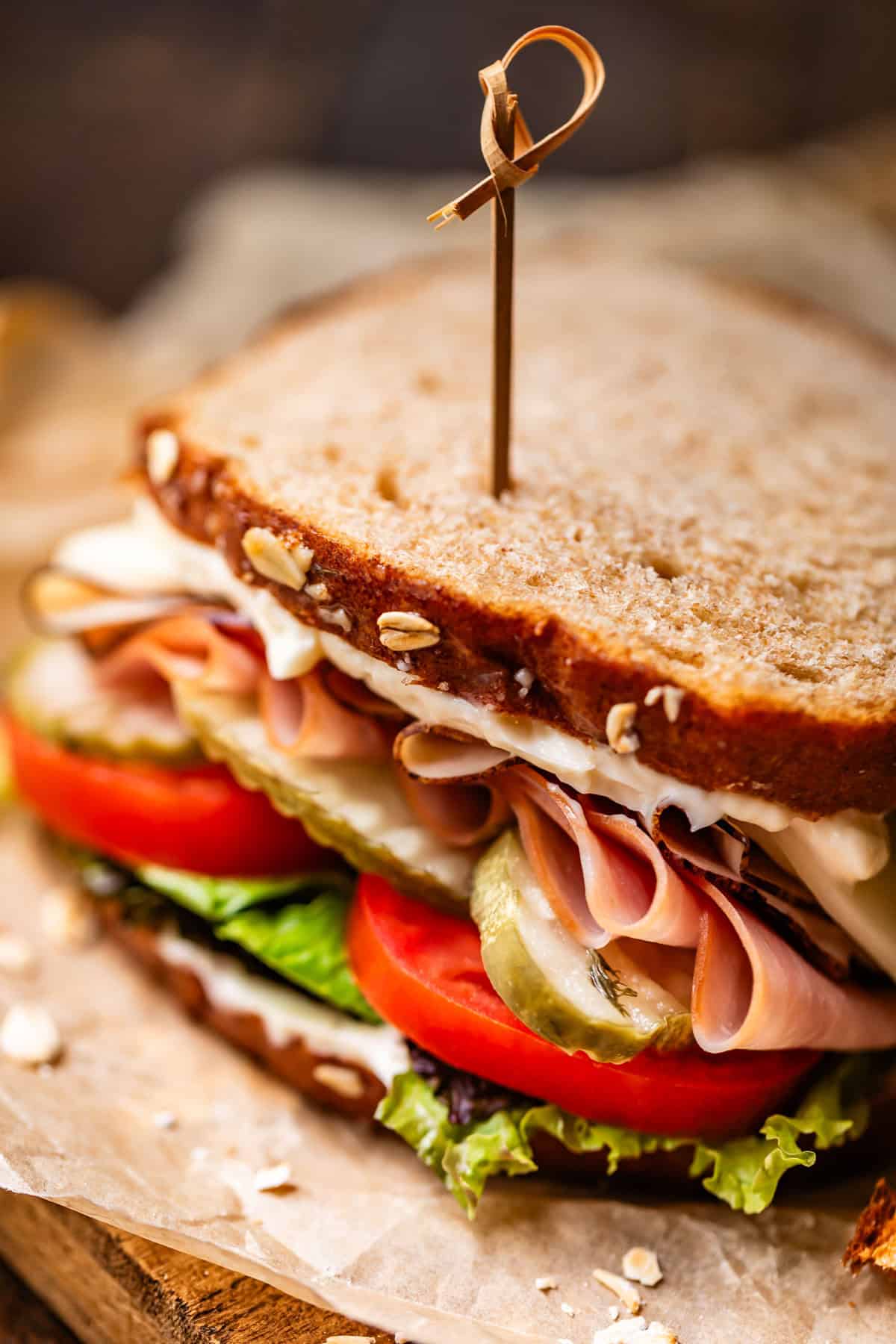 Close-up of a sandwich on whole grain bread, filled with lettuce, tomato slices, pickles, ham, cheese, and mayonnaise, secured with a wooden pick.