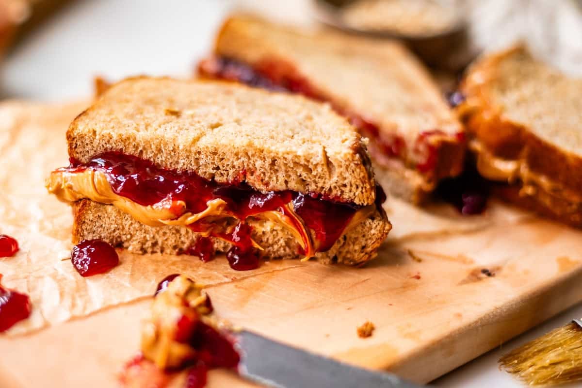 A close-up of a peanut butter and jelly sandwich cut in half on a wooden cutting board, with peanut butter and jelly oozing from the sides.