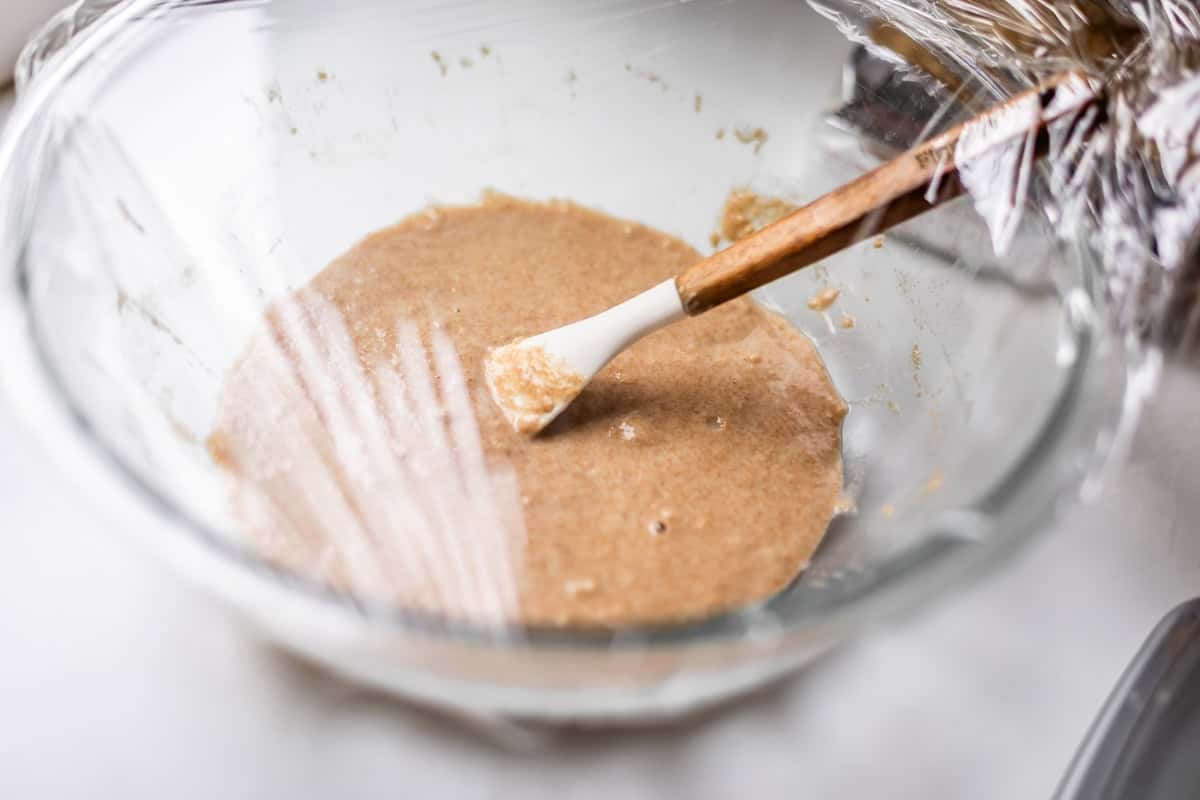 A glass bowl of brown batter is partially covered with plastic wrap. A white and wooden spatula rests inside the bowl. The bowl sits on a white surface.
