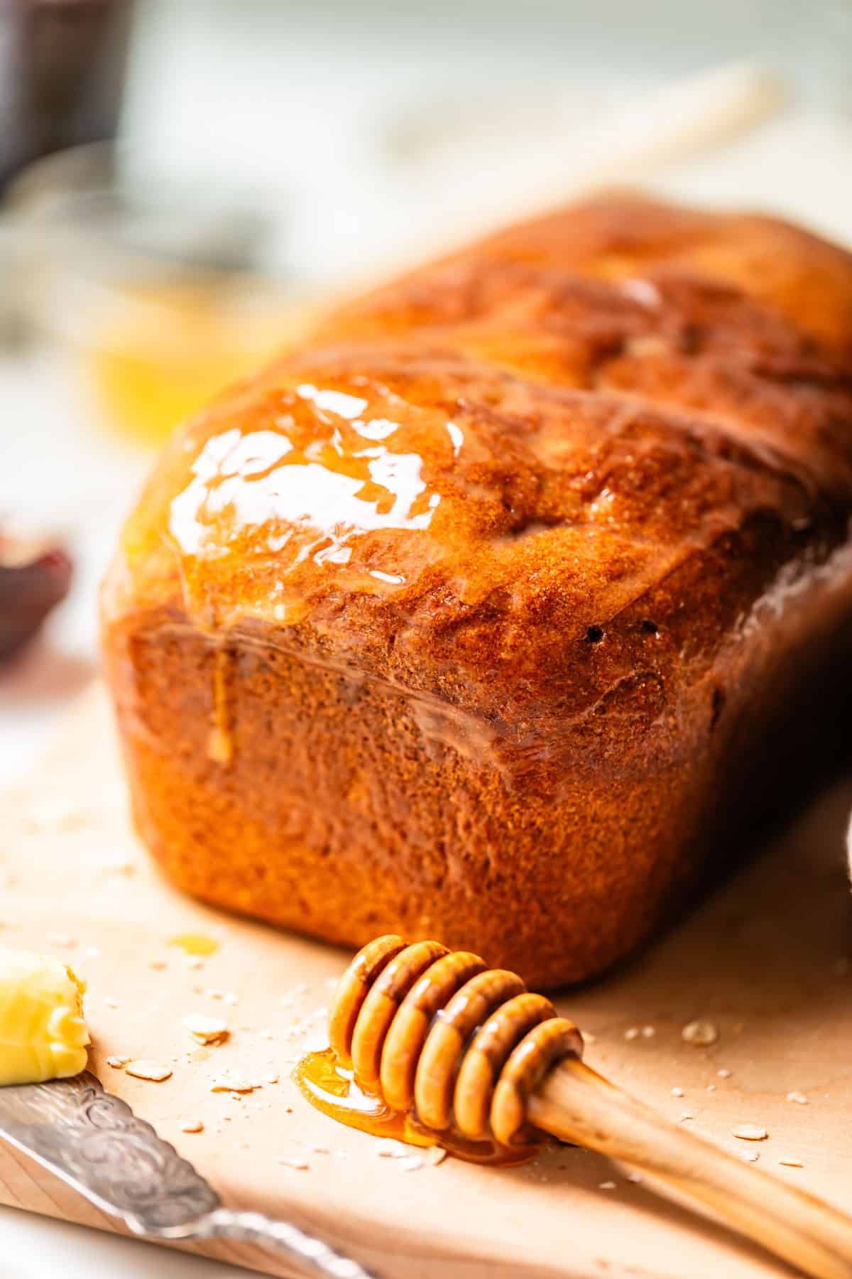 A loaf of bread covered with honey sits on a wooden board, with a honey dipper and a pat of butter nearby.