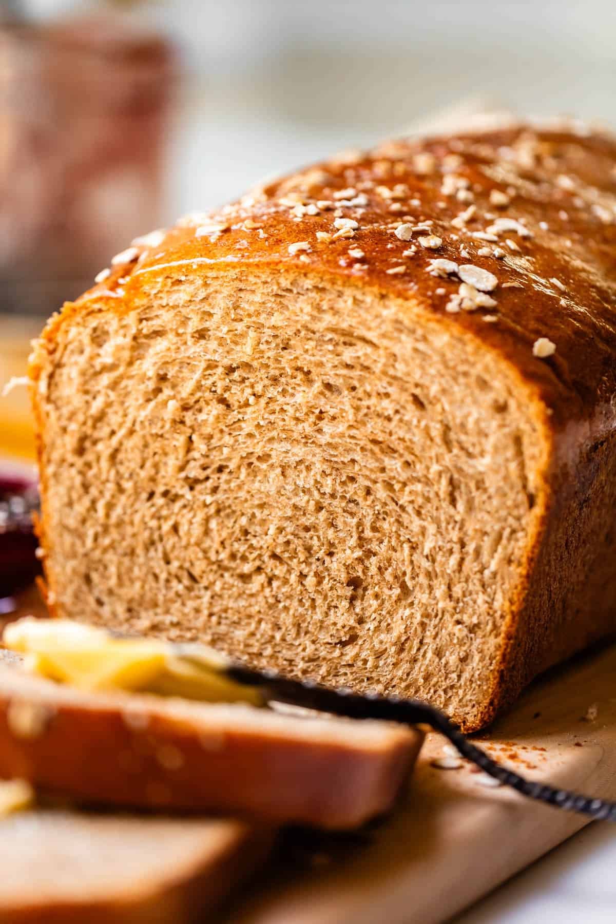 A close-up of a loaf of whole wheat bread on a wooden board, with two slices cut and some butter and jam visible in the foreground. Oats are sprinkled on top of the loaf.