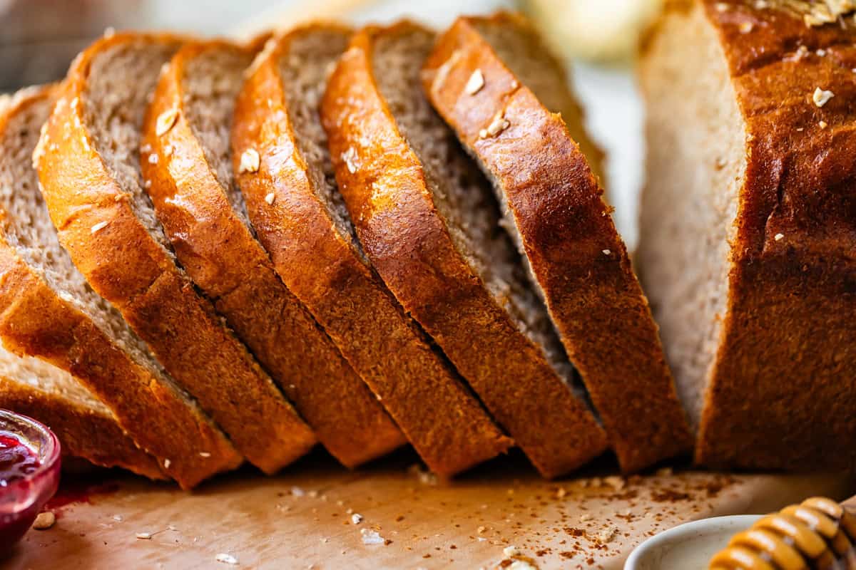 A close-up of several slices of freshly baked bread arranged in a row on a wooden surface, with a loaf in the background and a small dish of jam partially visible beside them.