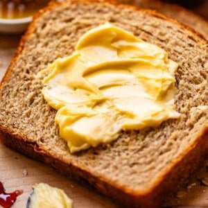 A slice of whole grain bread with creamy butter spread on top, surrounded by a honey dipper, jam, and a butter knife with more butter.