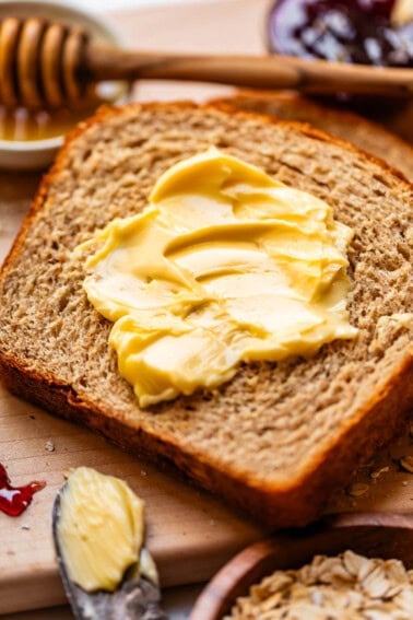 A slice of whole grain bread with creamy butter spread on top, surrounded by a honey dipper, jam, and a butter knife with more butter.