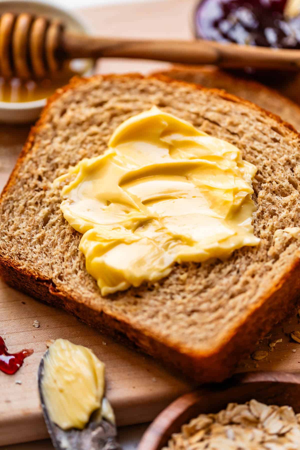 A slice of whole grain bread with creamy butter spread on top, surrounded by a honey dipper, jam, and a butter knife with more butter.