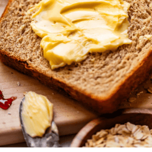 A slice of whole wheat bread with butter spread on top sits on a wooden surface near a spoon with more butter, oats, jam, and a honey dipper. Text below reads, Honey Whole Wheat Bread - The Food Charlatan..