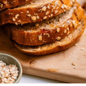 Sliced whole wheat honey bread on a wooden cutting board, topped with oats. A bowl of oats is nearby. Text at the bottom reads: Whole Wheat Honey Bread. The Food Charlatan..