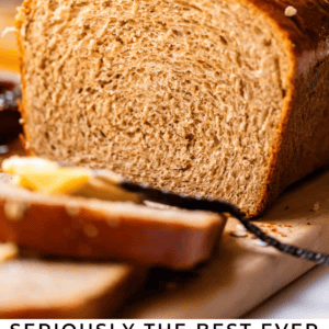 A close-up of a loaf of honey wheat bread on a cutting board with a slice cut. Butter and jam are on the side. Text reads: Seriously the Best Ever Honey Wheat Bread. The Food Charlatan..