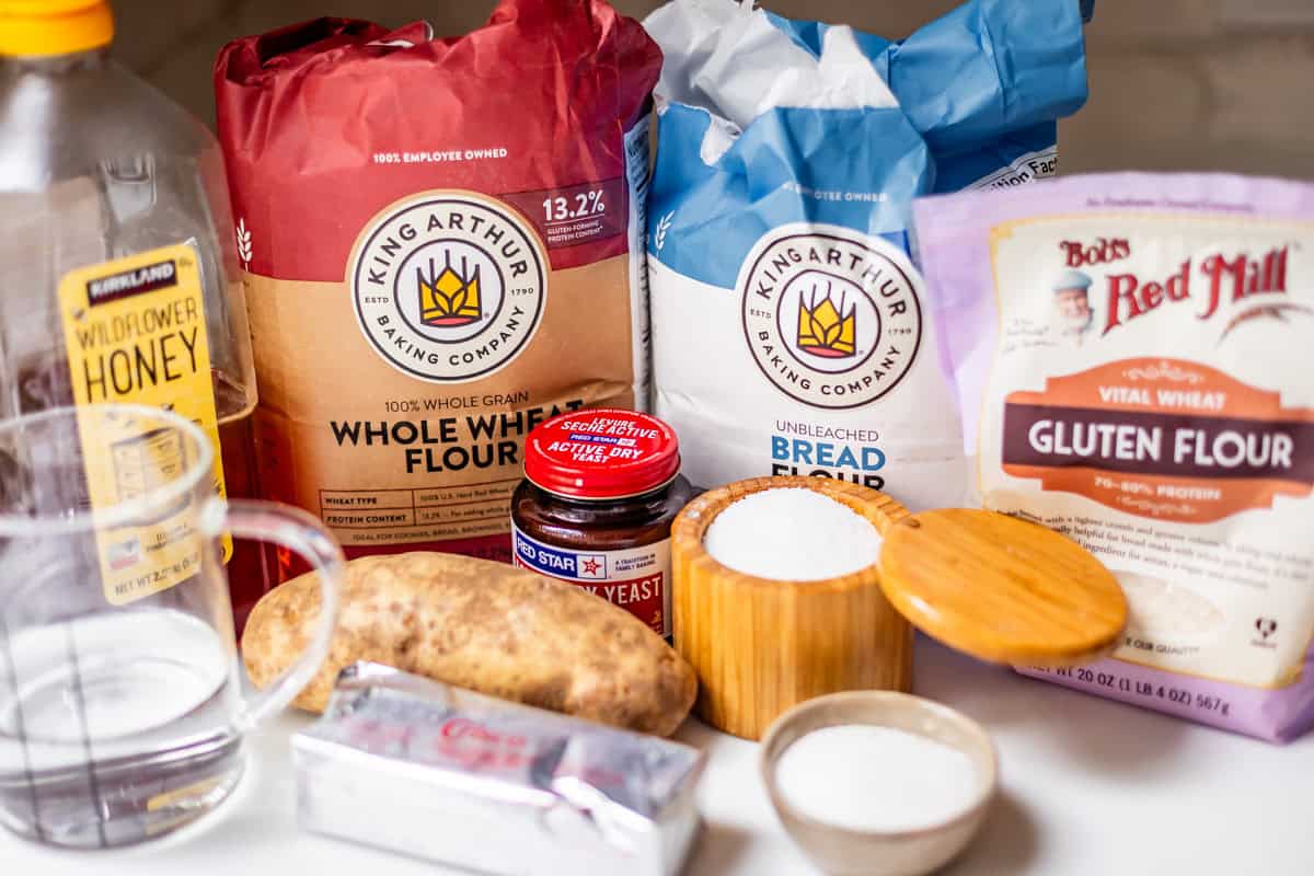 Various bread-making ingredients on a countertop, including flours, honey, yeast, butter, a potato, a glass measuring cup, and containers of salt and sugar.