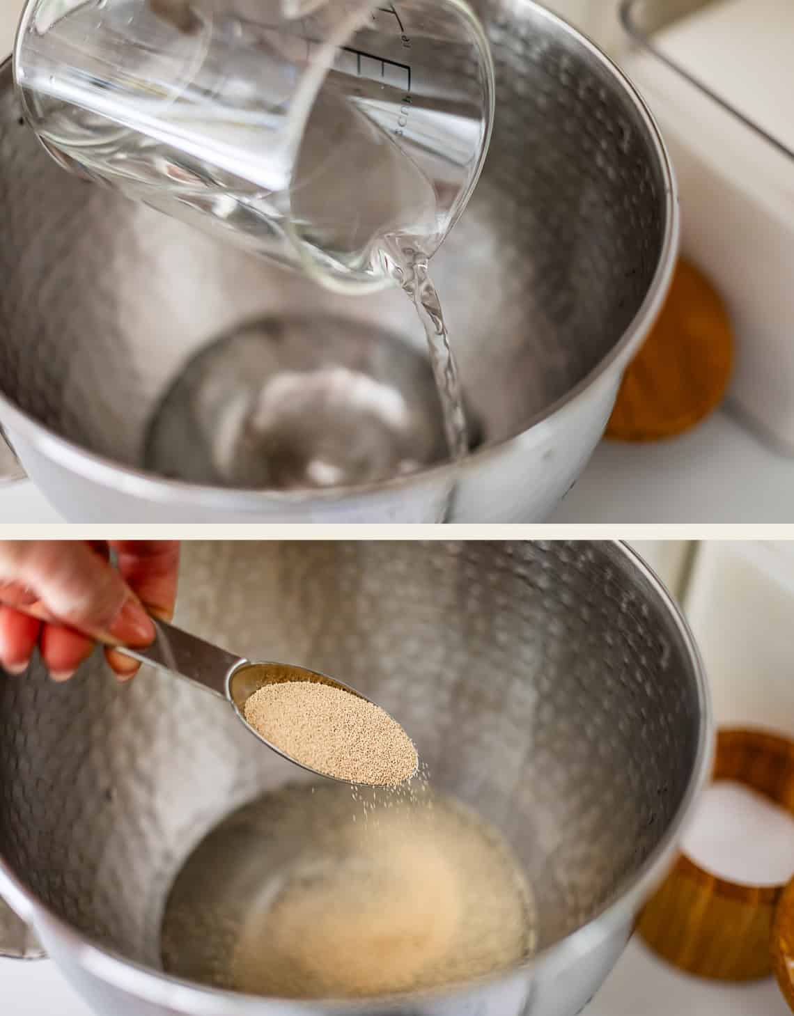 Water being poured from a measuring cup into a metal mixing bowl, followed by a hand using a measuring spoon to add dry yeast to the water.