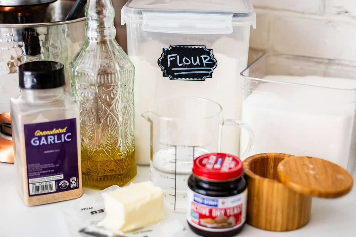 Baking ingredients on a counter, including a container labeled flour, garlic powder, oil, water, a stick of butter, active dry yeast, and a sugar container.