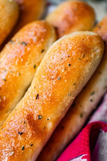 A close-up of golden-brown garlic breadsticks sprinkled with herbs, resting in a red and white cloth.
