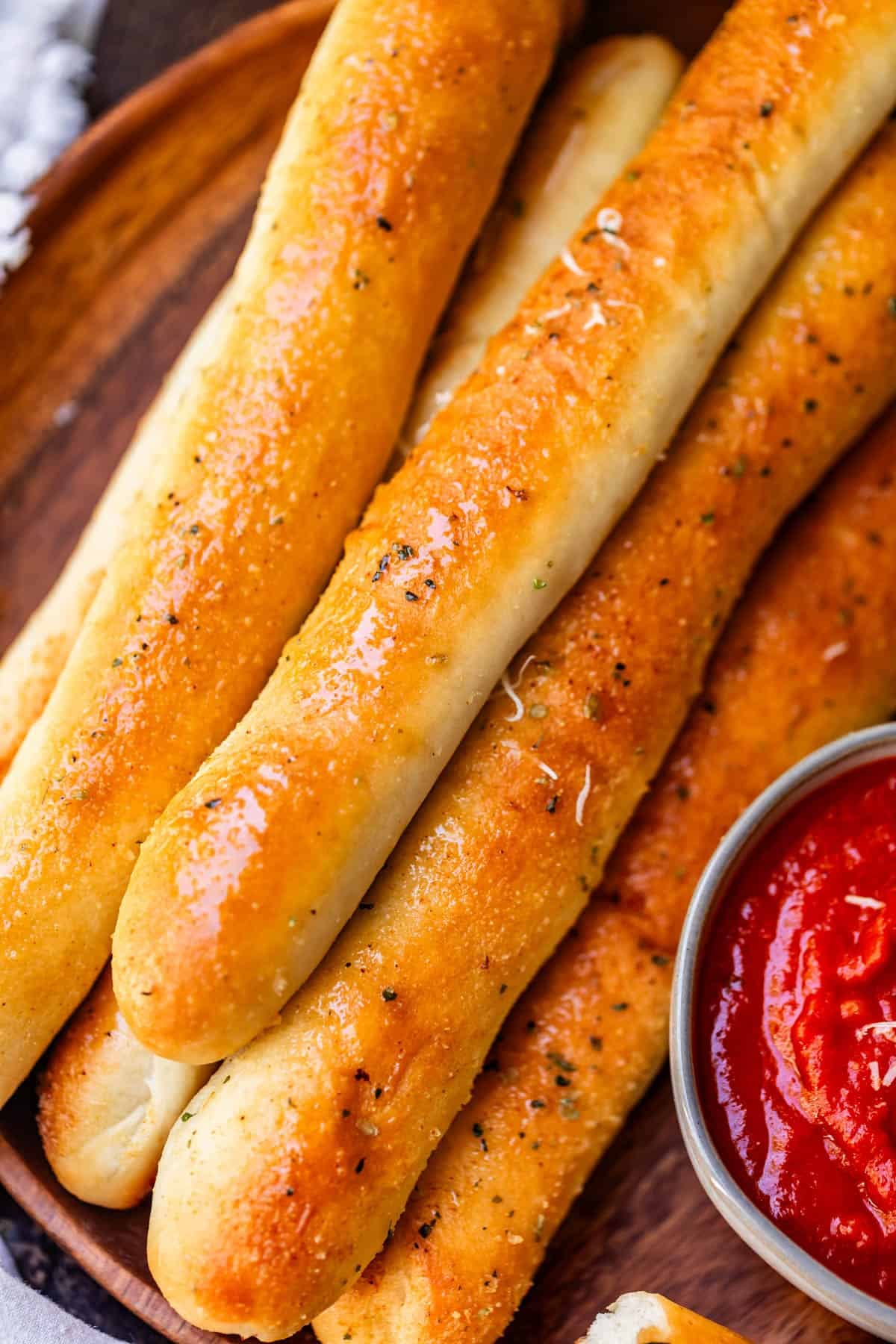 A pile of golden-brown garlic breadsticks sprinkled with herbs, served with a small bowl of marinara sauce.