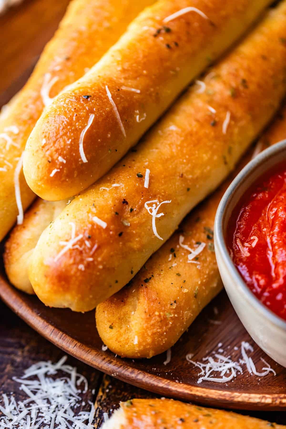 A wooden plate piled with golden-brown breadsticks sprinkled with herbs and cheese, served with a side of marinara sauce.