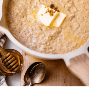 A pot of cooked oatmeal topped with a pat of melting butter and a sprinkle of brown sugar, with a honey dipper and spoons nearby.