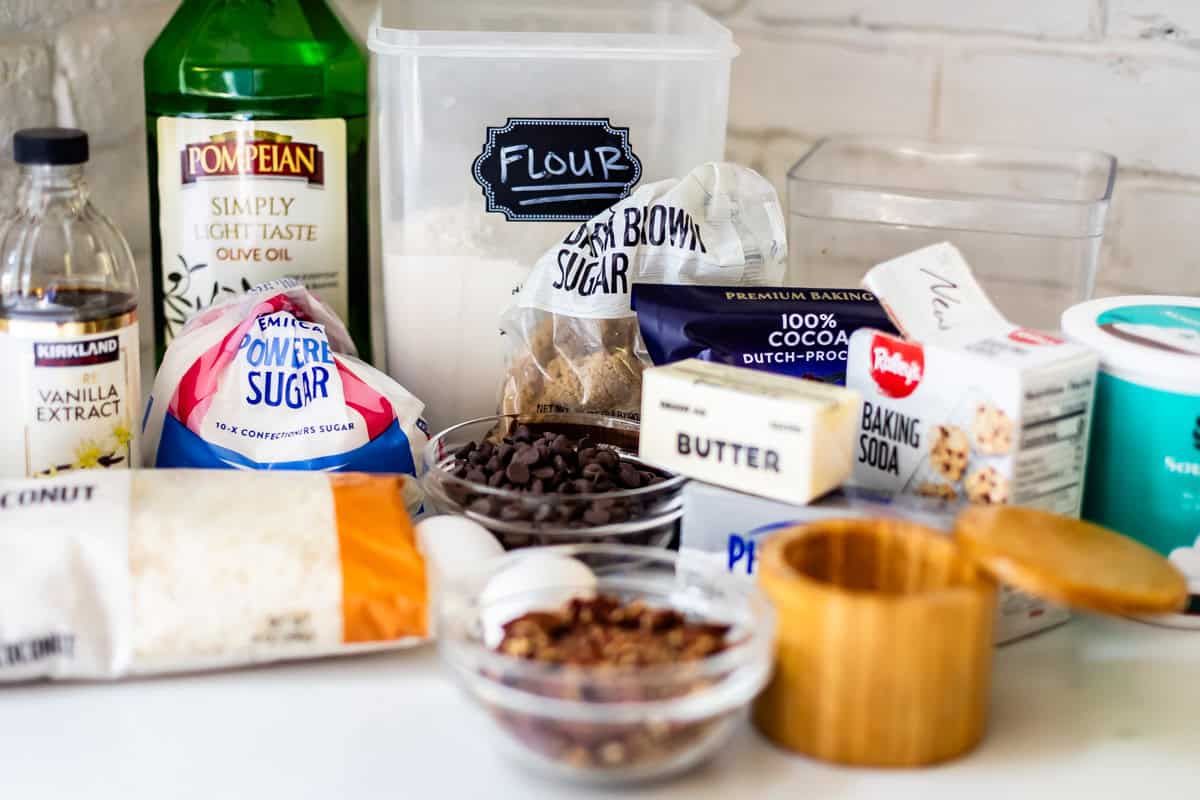 A kitchen counter filled with baking ingredients, including olive oil, vanilla extract, powdered sugar, flour, brown sugar, cocoa powder, butter, chocolate chips, baking soda, shredded coconut, eggs, nuts, and a salt cellar.