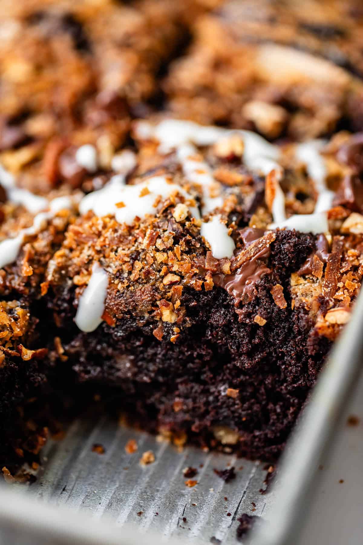 Chocolate cake in a baking pan topped with toasted coconut, nuts, chocolate chips, and a white glaze.
