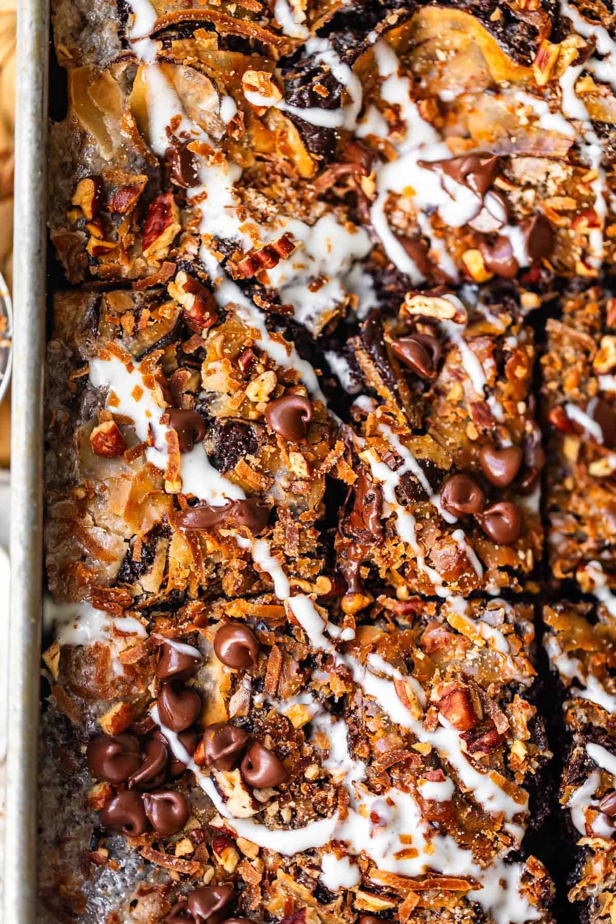 Sheet pan of chocolate bars topped with pecans, shredded coconut, chocolate chips, and a white icing drizzle.