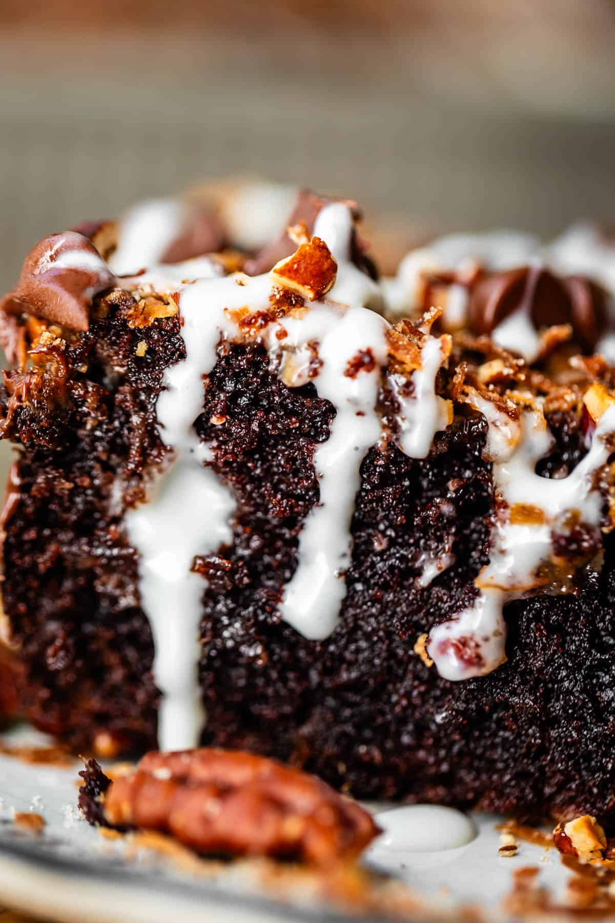 A close-up of a slice of dark chocolate cake drizzled with white icing, topped with chocolate chips and toasted nuts.