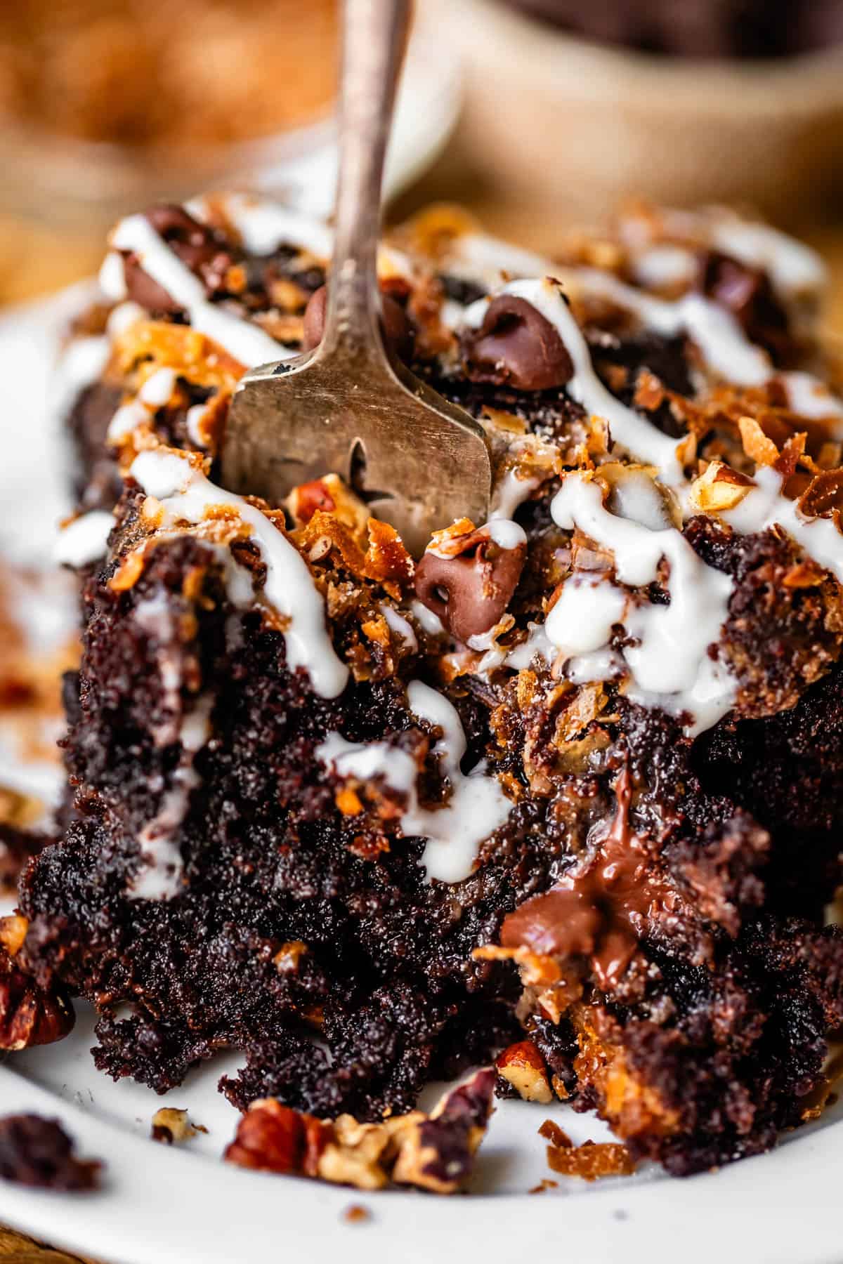A slice of chocolate cake topped with toasted coconut, chocolate chips, and white icing, being eaten with a silver fork.
