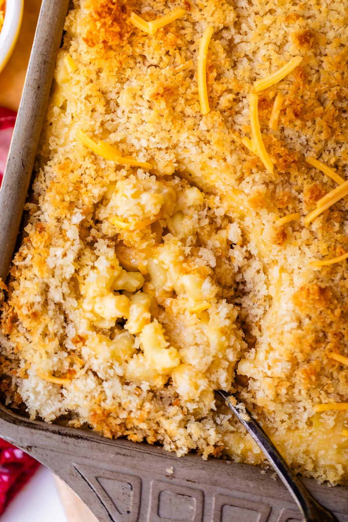 A metal baking pan filled with baked macaroni and cheese topped with breadcrumbs and shredded cheese, with a portion scooped out.